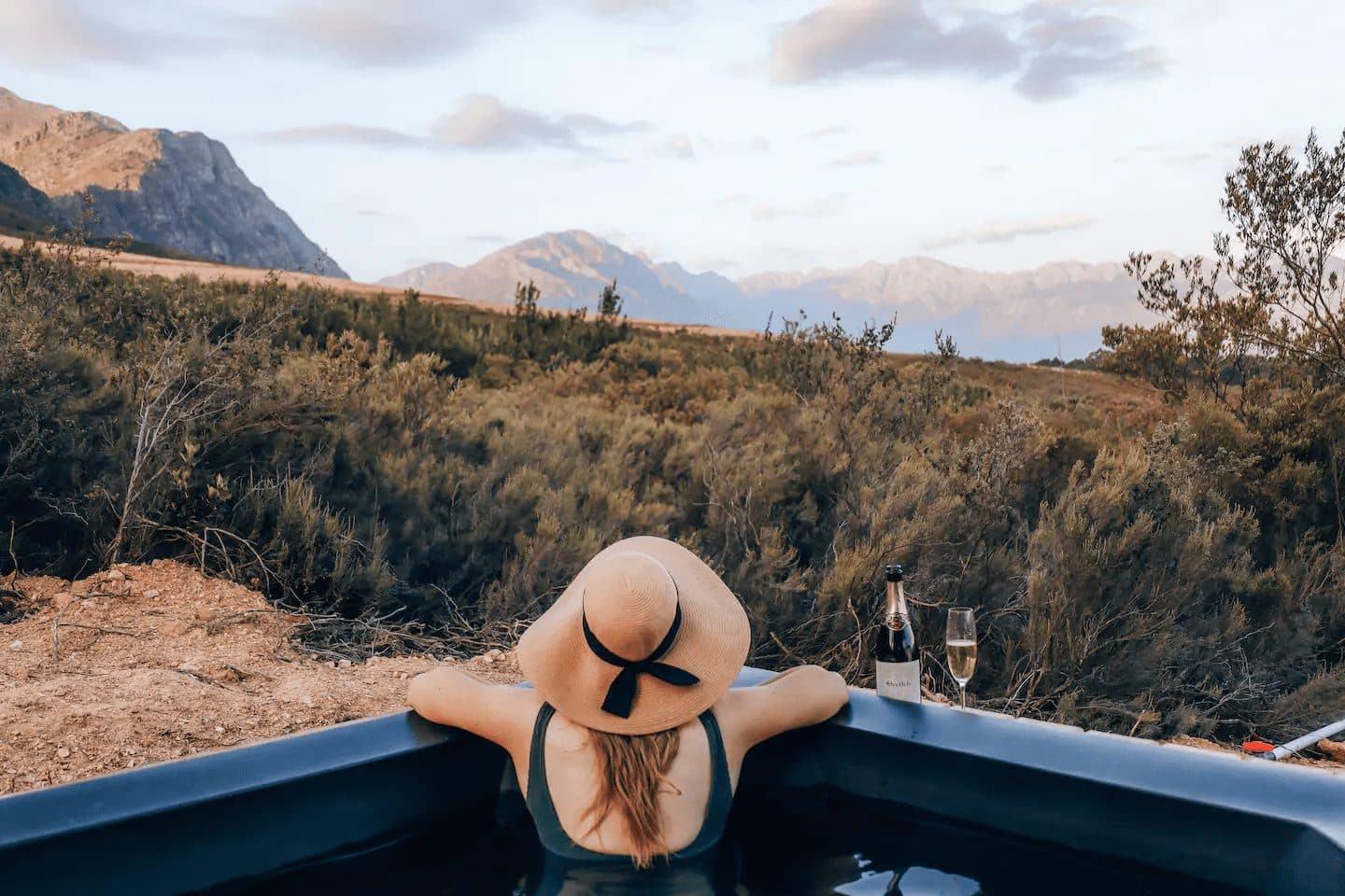 Radio Nowhere Cabin: Woman in hot tub with sunhat, champagne, mountains at dusk